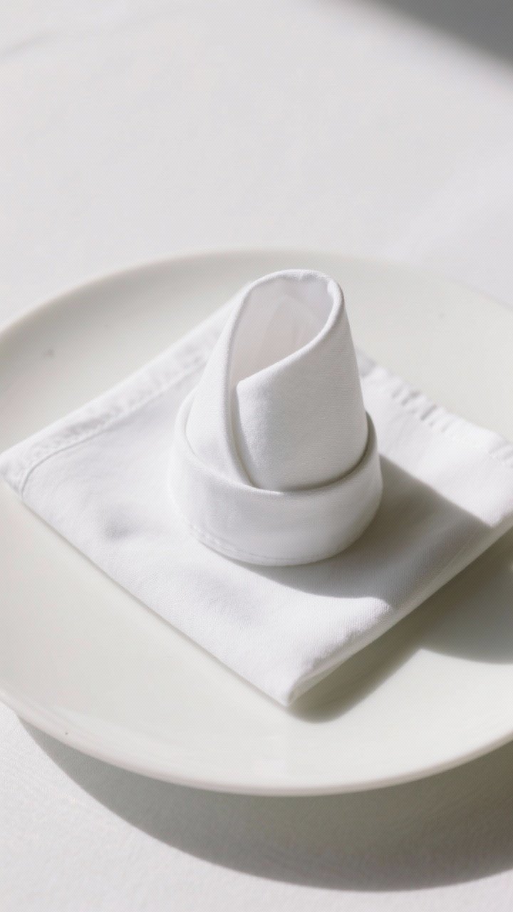 Overhead shot of a neatly folded Bishop’s Hat napkin on a white porcelain plate, using a single 18–20 inch square cloth napkin in crisp white linen, edges sharply defined with light starch; clean flat tabletop, soft natural side light, minimal props to emphasize the elegant butler-like form.